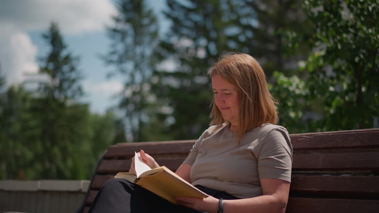 Une femme assise sur un banc en bois, lisant un livre à l’extérieur sous la lumière du soleil, avec une expression détendue et concentrée, tandis qu’une légère brise fait bouger les feuilles et que des voitures floues se déplacent en arrière-plan.
