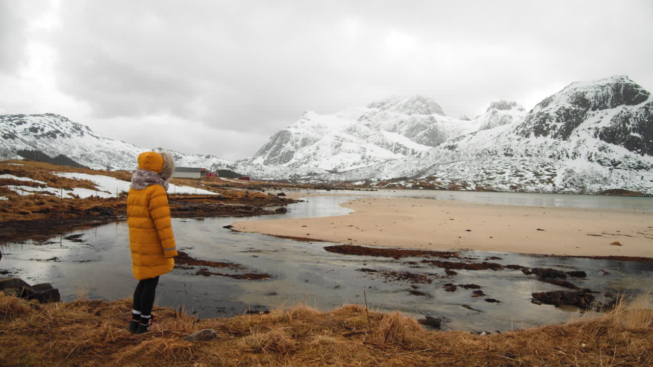 Person In Yellow Jacket Running Towards Edge Of Coast For Better View Of The Snowy Mountains In The Lofoten Islands, Norway - wide shot