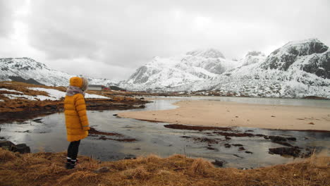 person in yellow jacket running towards edge of coast for better view of the snowy mountains in the lofoten islands, norway - wide shot