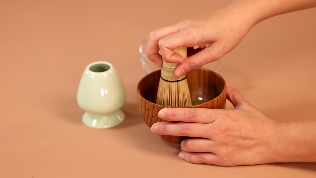Hands whisk matcha green tea with bamboo whisk in wooden bowl, minimal background, soft lighting
