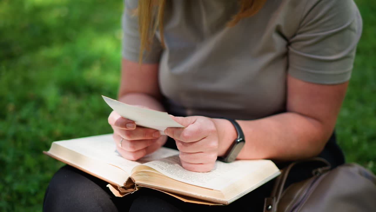 Close up of woman sitting outdoors holding photo found inside open book, looking curiously with emotional expression, peaceful sunlight and green background