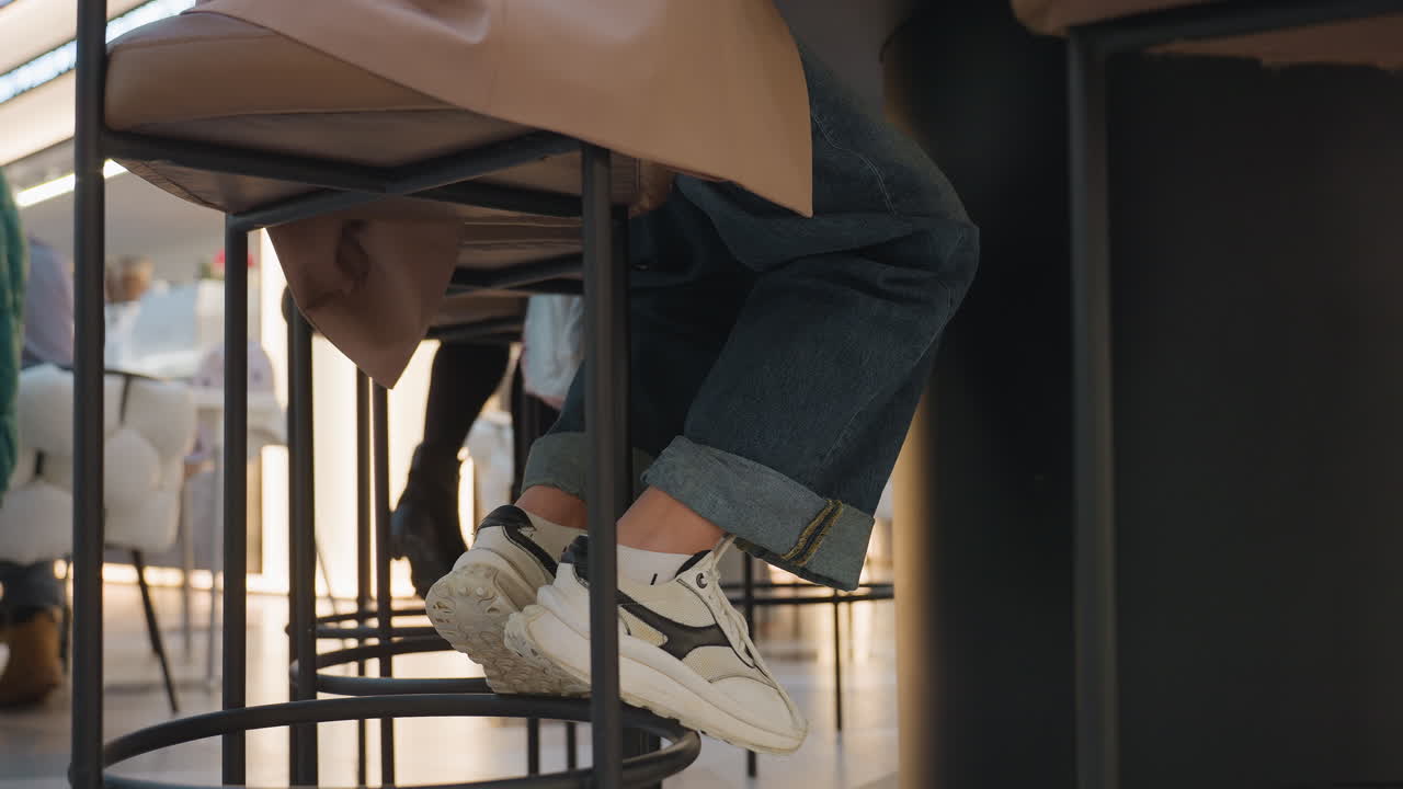 zapatillas bajo un taburete de bar, estudiante dibujando, bajos del vaquero enrollados, zapatos blancos con cordones apoyados en un reposapiés de metal, luz cálida del sol sobre suelo de madera, cuaderno de bocetos y café cerca, suaves golpecitos y relajación