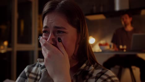 a brunette girl in a plaid shirt cries while watching a sad drama on tv and her brunette boyfriend in a gray t-shirt consoles the girl and hugs her in a modern studio apartment in the evening