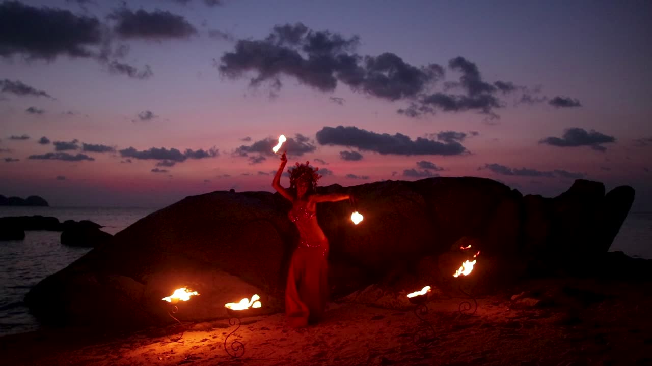 Slow motion of erotic and sensual belly dancer performing figure 8 move with fire palm torch props, on the beach during golden hour