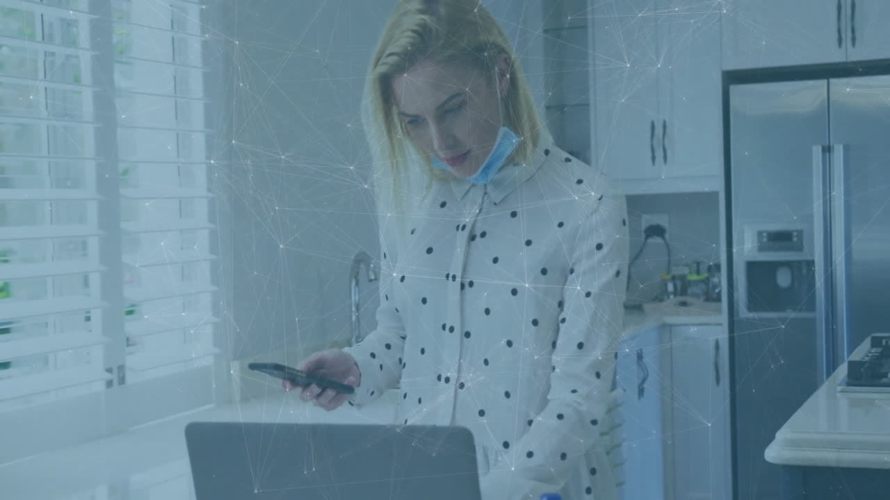 Woman standing at kitchen counter, holding smartphone, viewing laptop with animated business graphs
