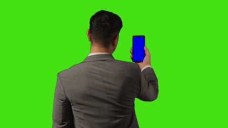 vista trasera sobre el hombro de un hombre de negocios con un teléfono móvil de pantalla azul de pie contra un fondo de pantalla verde
