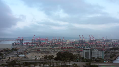 Grúas-De-Carga-Y-Contenedores-De-Envío-En-El-Puerto-De-La-Terminal-Industrial-En-Long-Beach,-California,-Con-Tráfico-Acelerado-De-La-Ciudad---Vista-Aérea