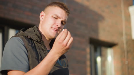 young male student holding cigarette looks conflicted under sunlight then discards cigarette with visible determination, brick wall and window in background