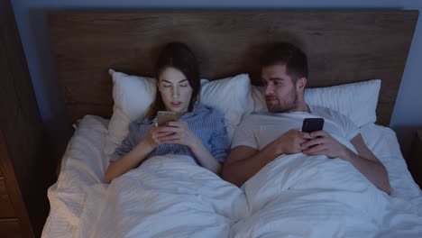 Vista-Superior-De-La-Pareja-Caucásica-Recién-Casada-Después-De-Una-Pelea-Tirada-En-La-Cama-Por-La-Noche-Y-Escribiendo-En-Los-Teléfonos-Inteligentes,-Luego-Se-Miran-Entre-Sí