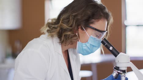 Female-teacher-wearing-face-mask-and-protective-glasses-using-microscope-in-laboratory