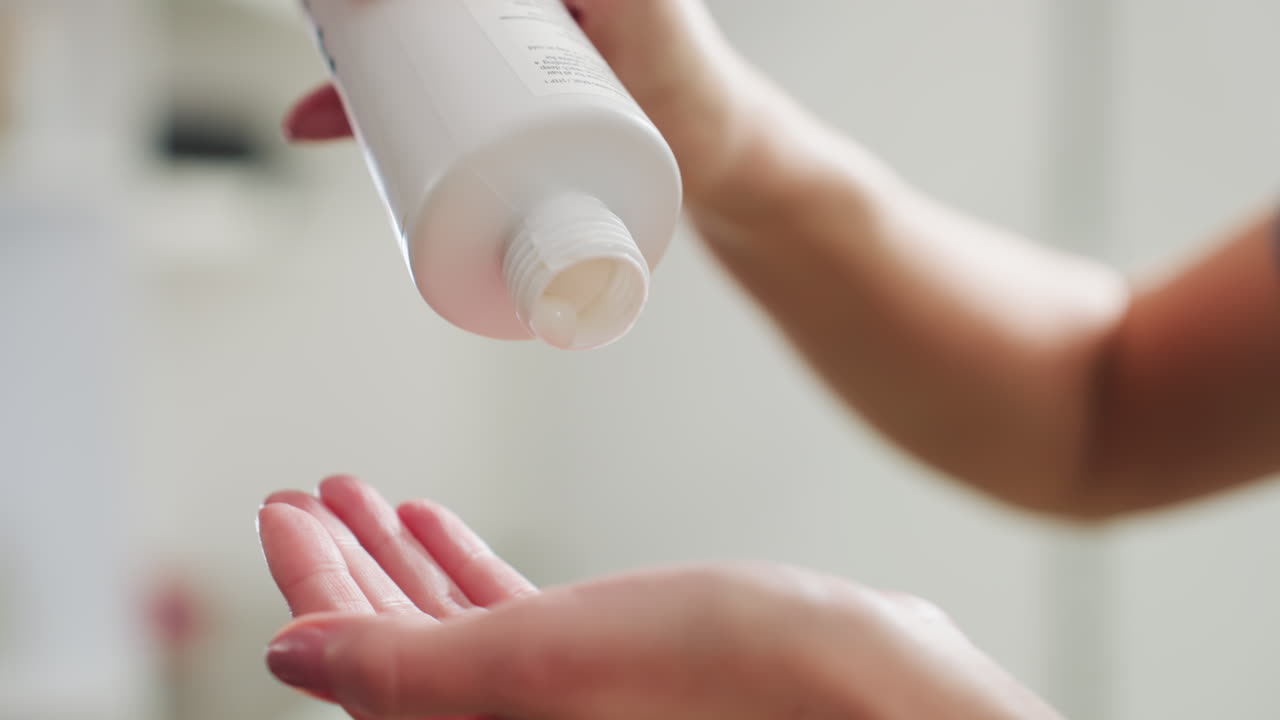 하얀 로션이 손바닥에 쏟아지는 모습, 크림 타입의 모이스처라이저를 짜서 받는 손의 클로즈업, 부드럽게 흐르는 질감, 은은한 욕실 조명, 수분 공급과 촉각적인 부드러움 강조