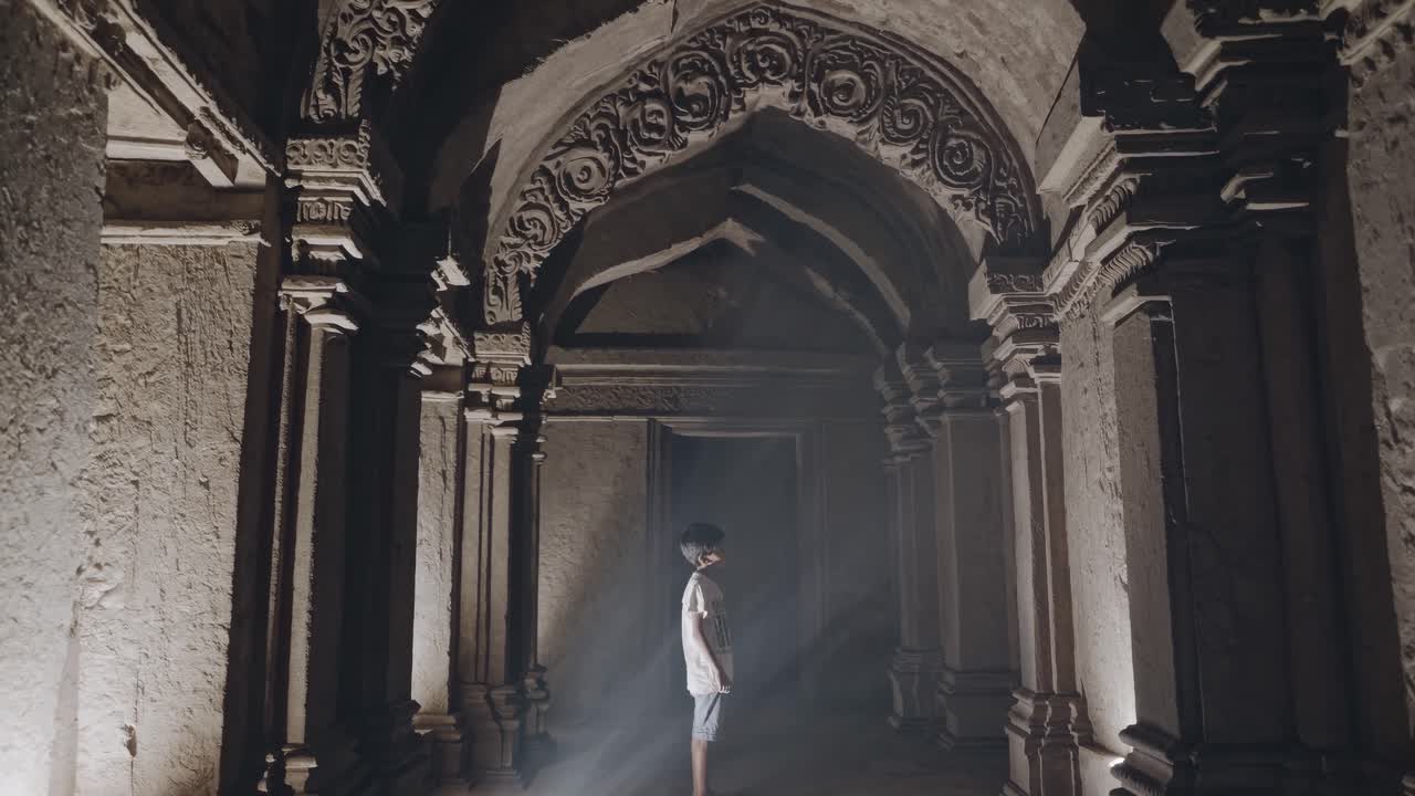 Sunbeam illuminating a young boy standing in a mystical hallway adorned with ornate columns and decorated arches, revealing the wonders of an ancient temple in Asia