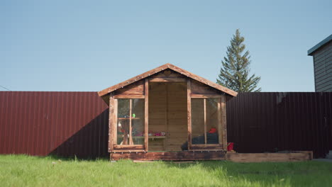 wooden kids playhouse with transparent glass panels sits on vibrant green grass, accompanied by sandbox, bordered by tall brown fence with pine tree and blue sky forming peaceful backdrop