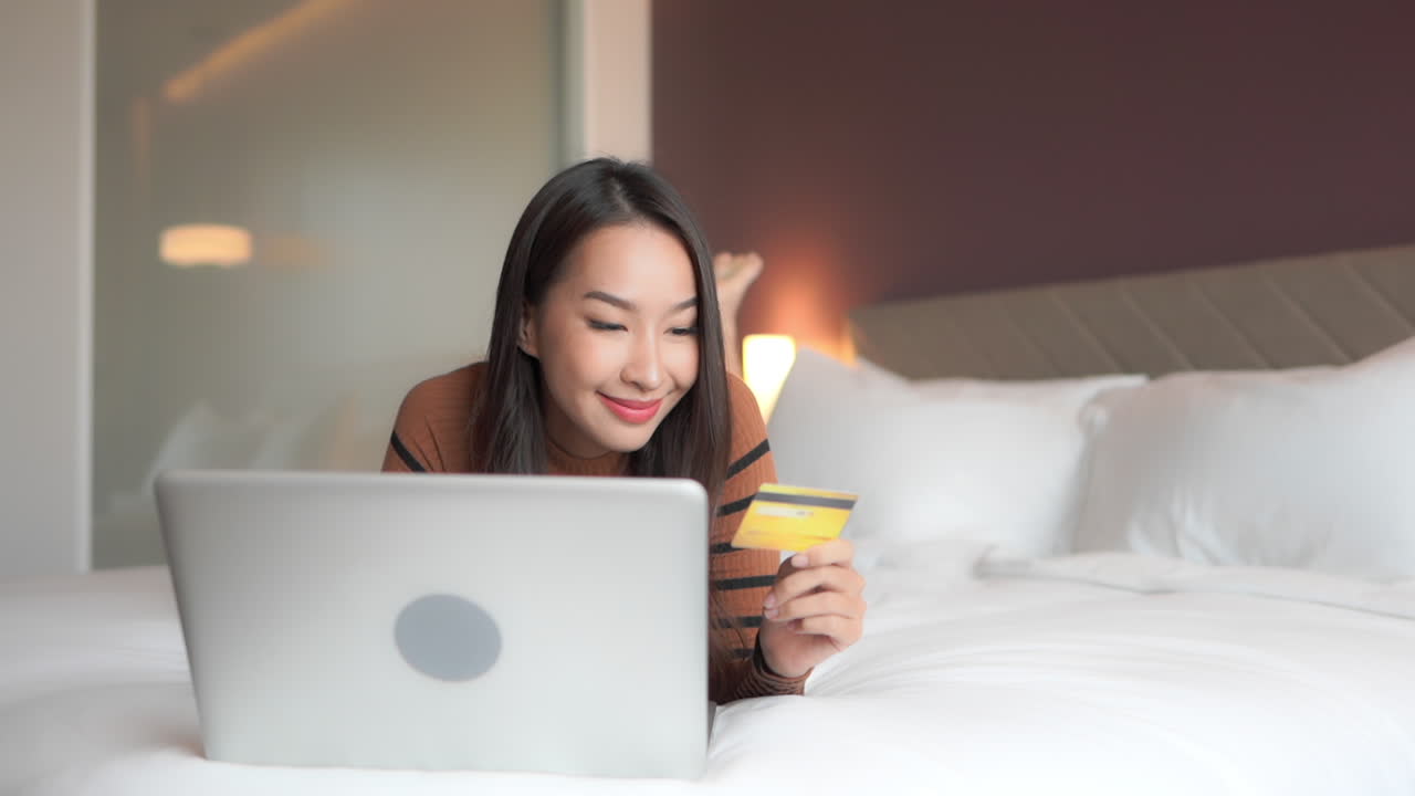 En ung, smuk kvinde ligger på maven og indtaster sine kreditkortoplysninger på sin laptop. Handel