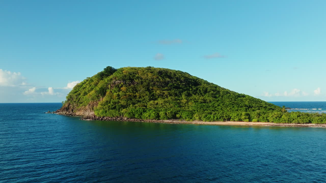 FPV drone flies laterally beside a small lush island off the coast of Guadeloupe, showing dense green vegetation, blue sea, and clear tropical sky