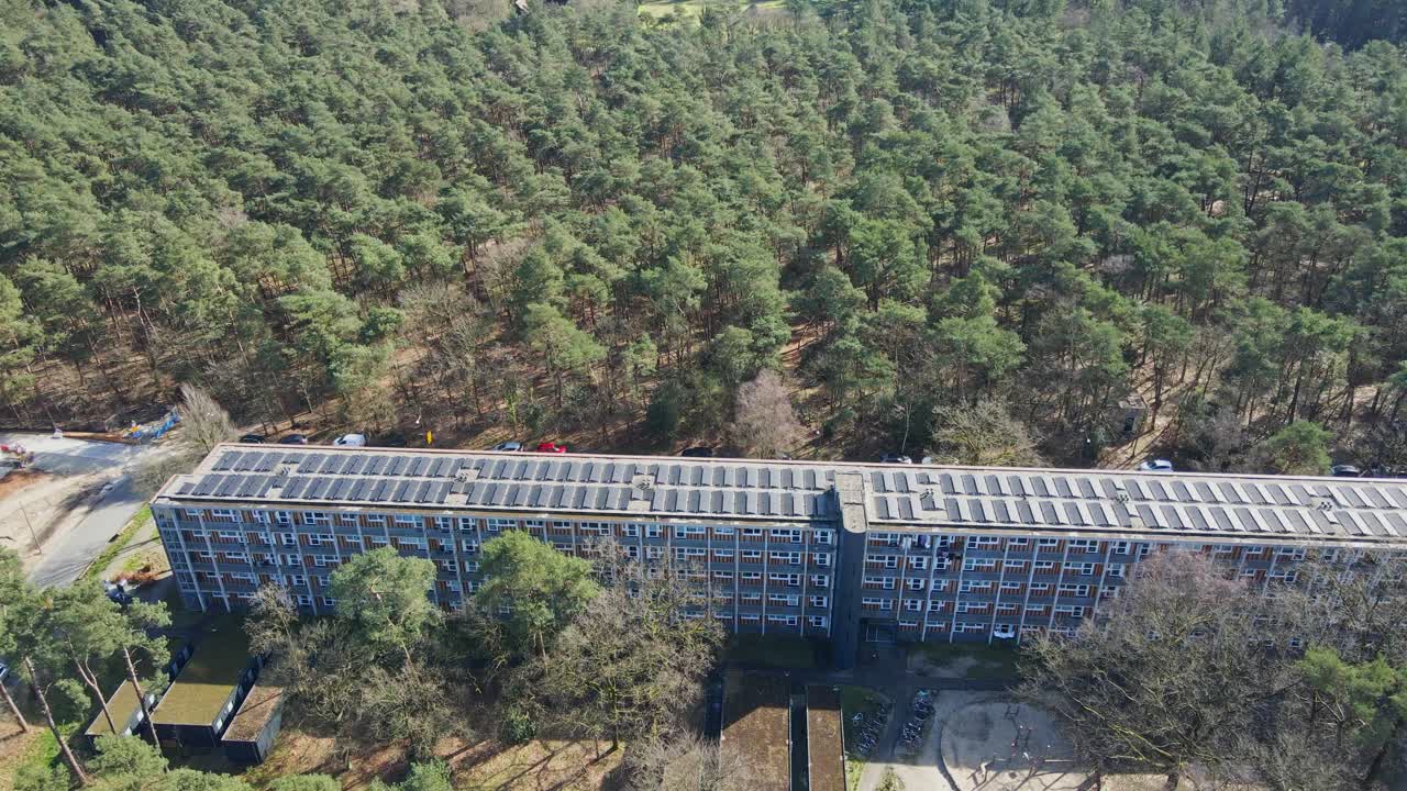 Aerial of modernized apartment building with photovoltaic solar panels on rooftop surrounded by a beautiful green forest on a sunny day