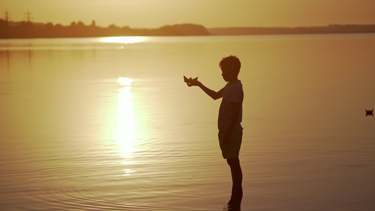 Boy with a Paper Ship is standing in the river. Origami Paper Boat in kid's hands. Beautiful Waves Ripple Background. Travel and nature Concept. Video at sunset.