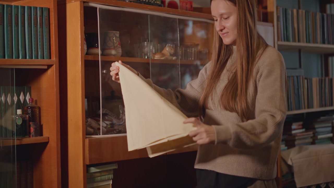 Kaukasische Frau entfaltet ein Erbstück-Tuch. Gemütliche Studienszene mit Holzschrank, Bücherregal im Hintergrund, sanftem Pullover, vorsichtigen Händen, die Porzellan- und Glassammlerstücke enthüllen, in warmem goldenem Licht.