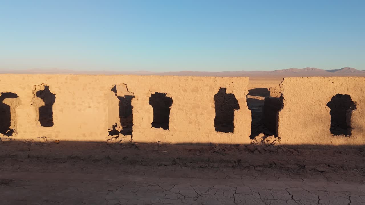 Side drone shot of an adobe facade in a deserted nitrate town at golden hour. Sunlight highlights the decay of a forgotten era in the vast Atacama Desert.