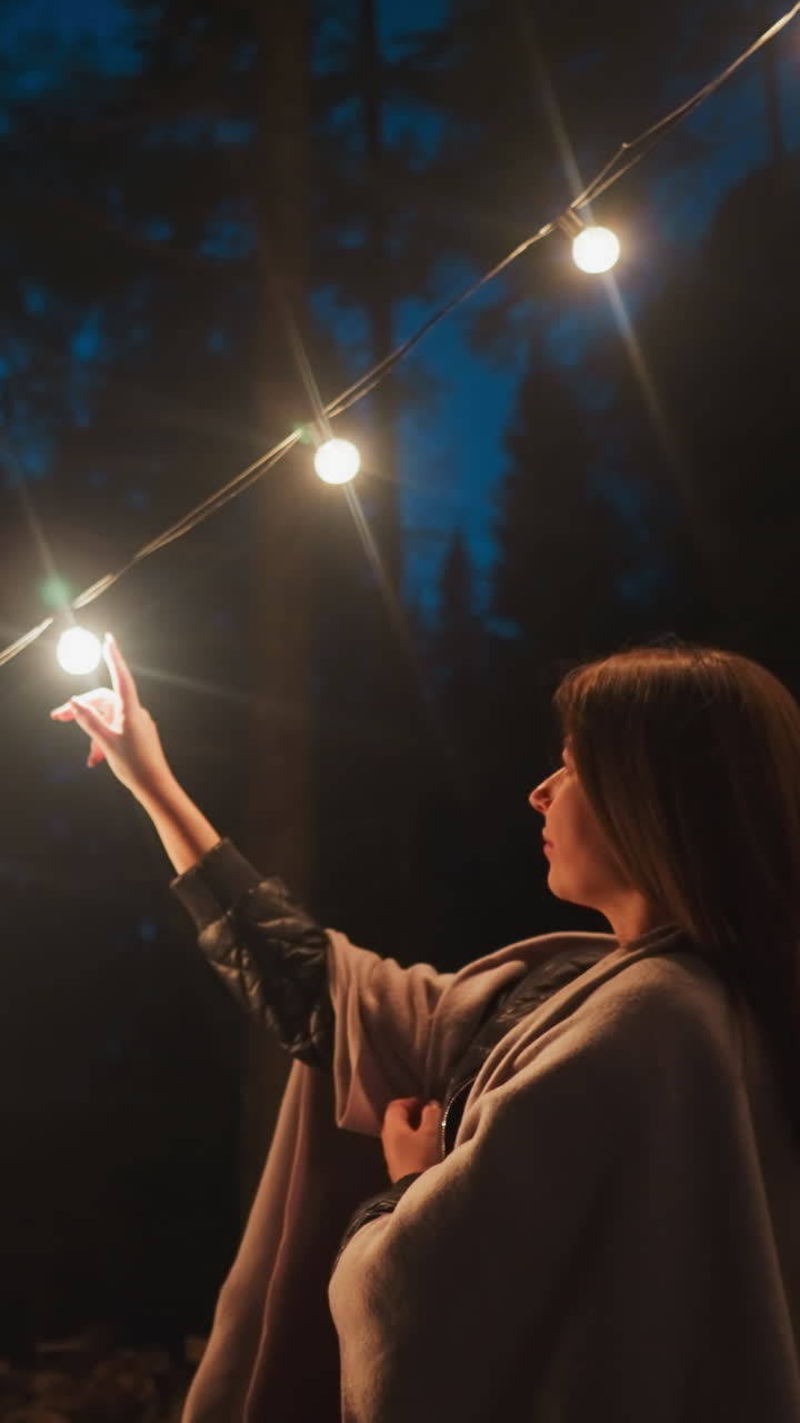 mujer acaricia una cadena de bombillas luminosas. el brillo de las luces suspendidas adorna los árboles que rodean la tienda de campamento a medida que cae la oscuridad. atmósfera encantadora