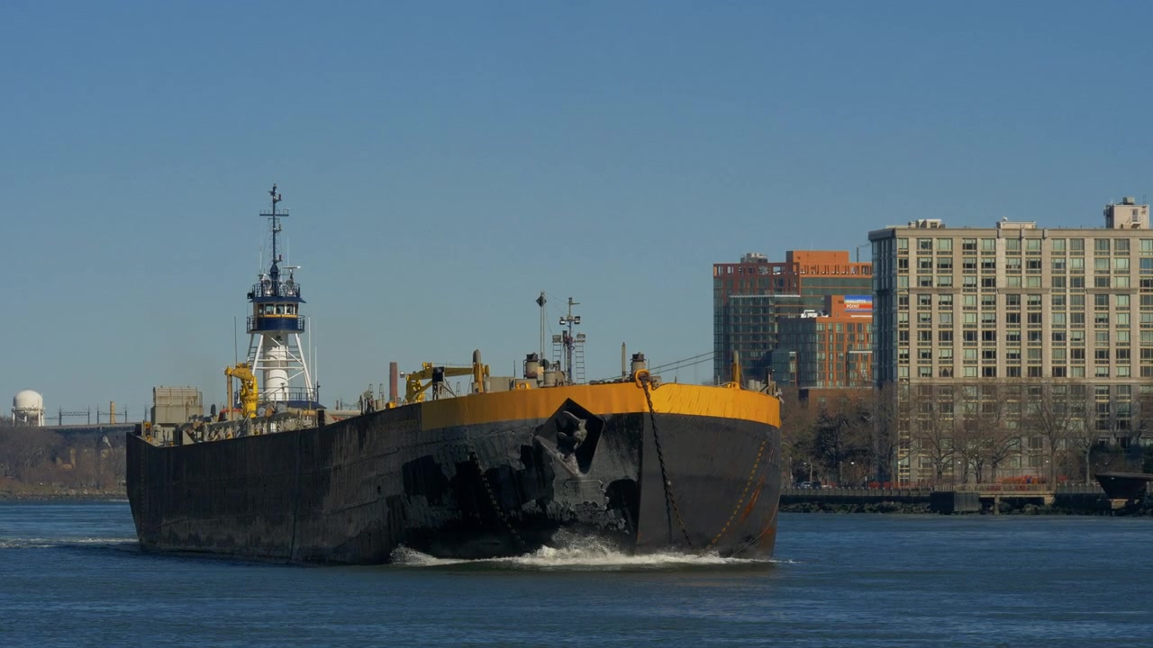 Vista ao nível do rio de uma grande barcaça de carga preta navegando no East River, passando pelos edifícios à beira-mar de Manhattan, com o apoio de um rebocador, água com ondulações e céu azul claro de inverno.