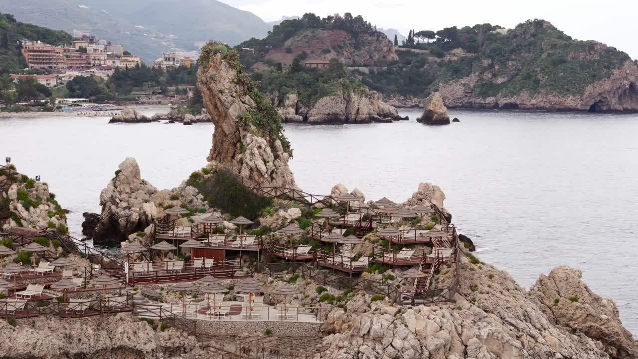 Aerial drone shot panning right of a fancy hotel on the rocks, revealing Isola Bella in the background, in Taormina, Sicily, Italy (Taormina, Sicilia, Italia)
