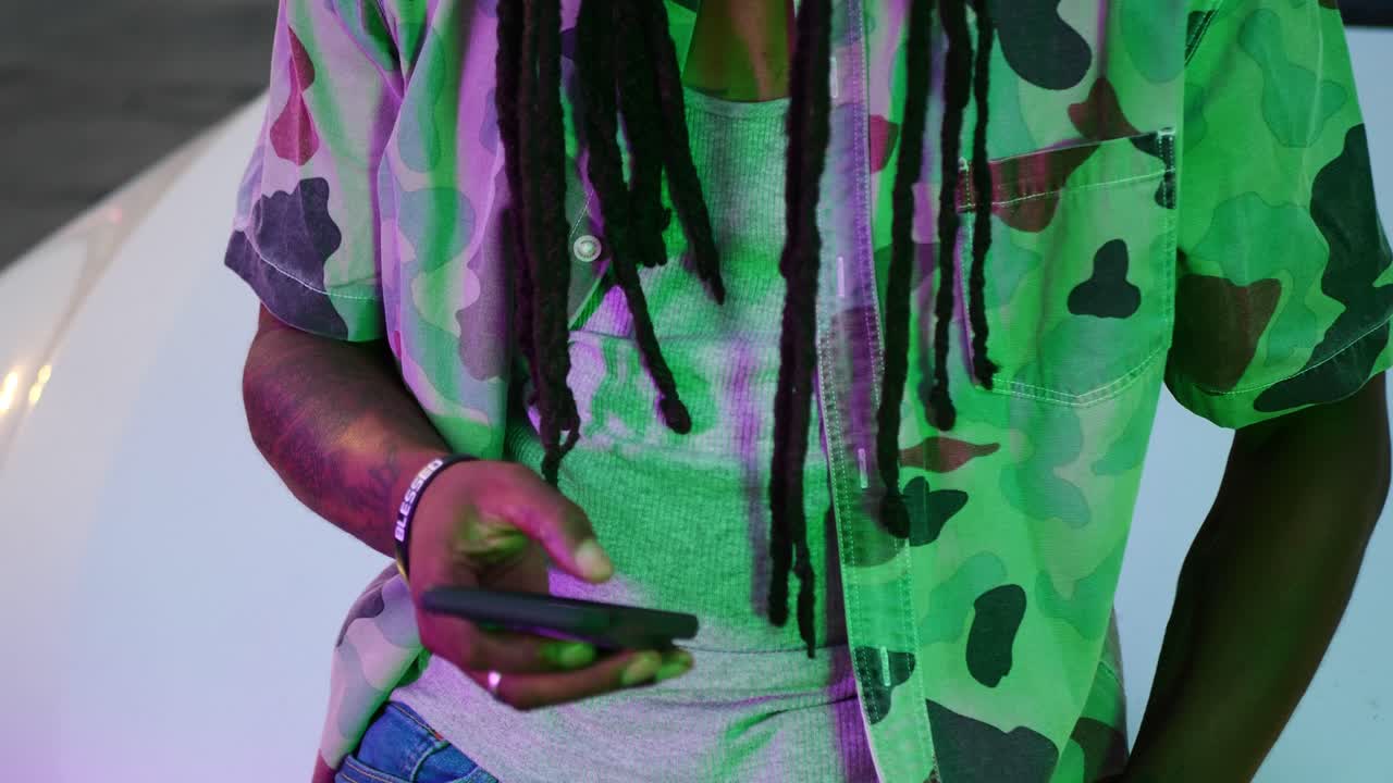 hombre afroamericano con rastas sentado en el auto y saca el teléfono para enviar mensajes de texto, luego hace muecas a la cámara