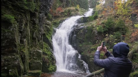 person taking photo of waterfall in autumn forest