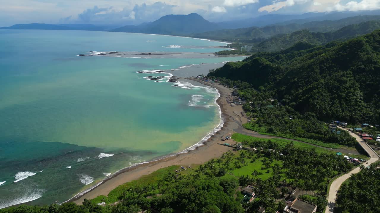 Tilt‑up aerial in Dingalan Aurora, lifting from the coastline to reveal drifting clouds above and a distant mountain rising on the horizon, framed by lush greenery and turquoise sea