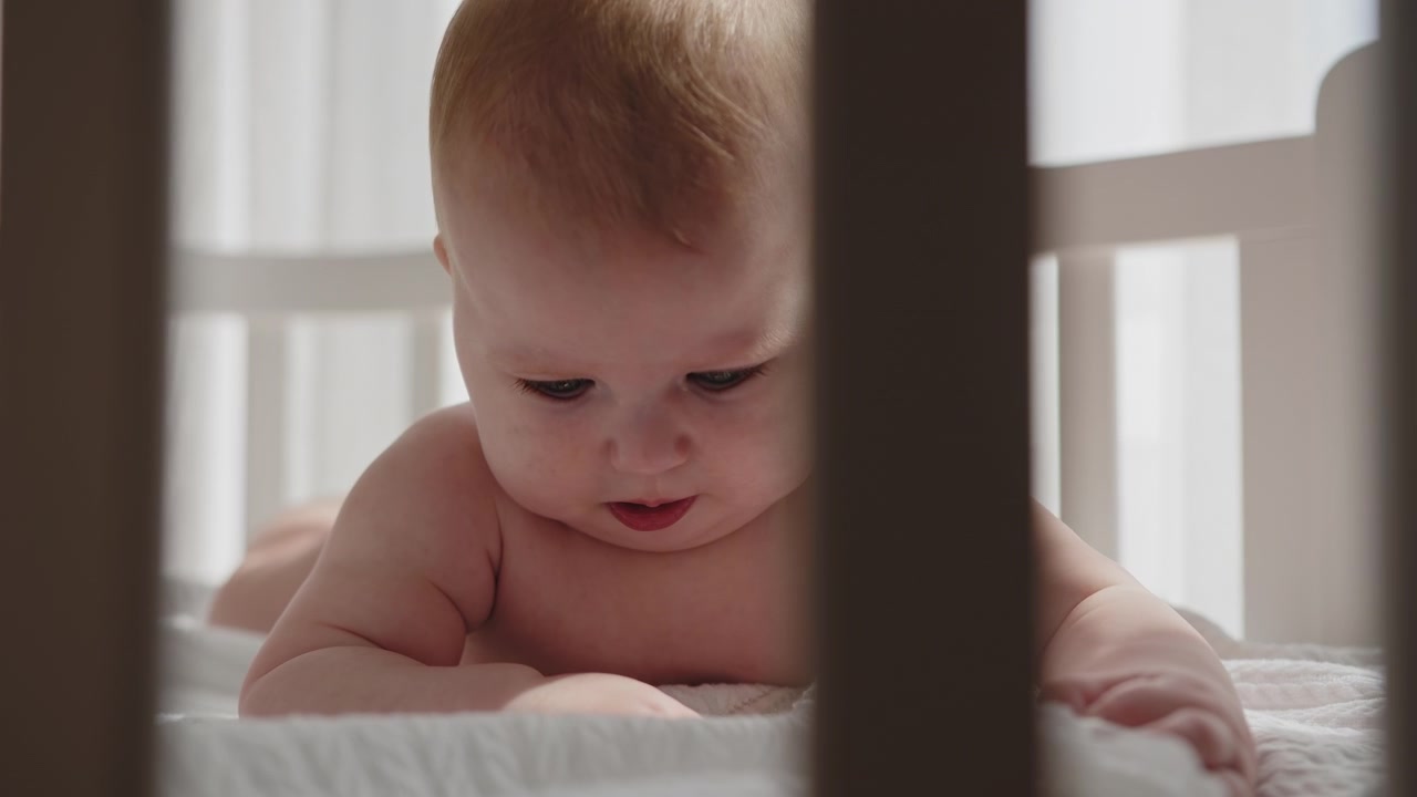 Closeup of infant exploring crib on soft white bedding with natural light filtering through bars a