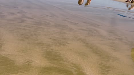 Nahaufnahme-Eines-Spaziergangs-Auf-Nassem-Sand-Mit-Reflexionen-Von-Menschen-An-Einem-Sonnigen-Tag