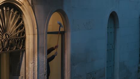 Person-cleaning-window-of-a-historic-building-in-Venice-at-dusk,-framed-by-arched-windows