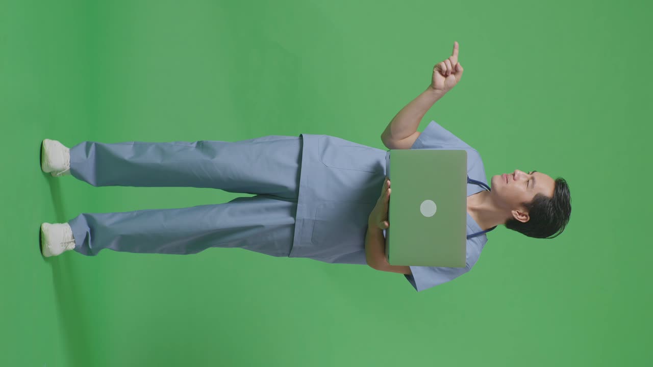 cuerpo lleno de médico masculino asiático con estetoscopio sosteniendo una computadora portátil, apuntando a un lado, y sonriendo en el fondo de la pantalla verde en el hospital