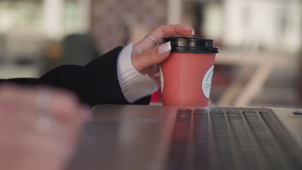 노트북 근처의 커피 컵을 부드럽게 돌리는 여자의 손의 클로즈업, 반지와 함께 그녀의 매니큐어 손가락에 초점을 맞추고, 의자와 함께 흐릿한 배경과 편안한 작업 환경