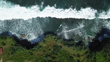 Vista-Aérea-De-Enormes-Olas-De-Mar-Que-Se-Estrellan-Contra-Acantilados-Cubiertos-De-Vegetación-Durante-La-Luz-Del-Sol