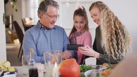 Video-Einer-Glücklichen-Kaukasischen-Mutter,-Tochter-Und-Großvater,-Die-Am-Esstisch-Auf-Ihr-Smartphone-Schauen