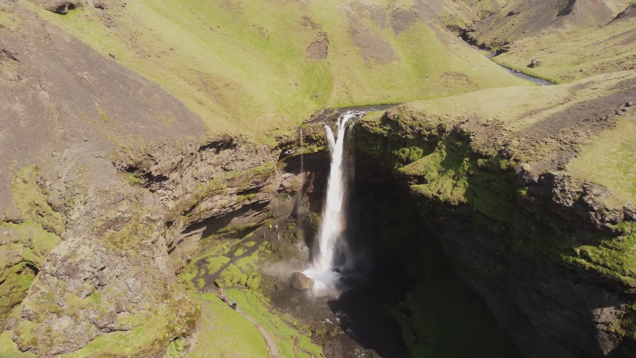 empuje hacia adentro y hacia abajo un disparo de drone de una hermosa cascada en islandia en un día soleado con pájaros volando y un pequeño arco iris frente a los acantilados y rocas verdes cubiertos de musgo