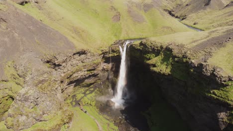 Push-in-and-down-drone-shot-of-a-beautiful-waterfall-in-Iceland-on-a-sunny-day-with-birds-flying-and-a-small-rainbow-in-front-of-the-mossy-green-cliffs-and-rocks