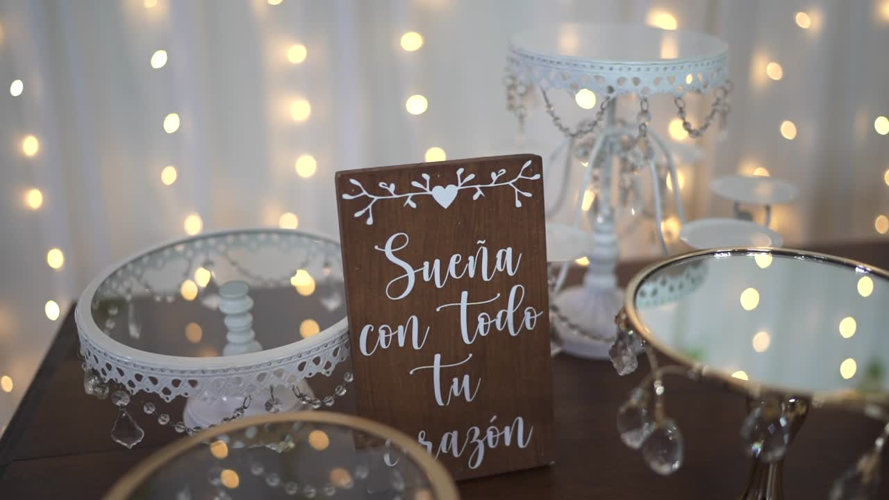 Table de mariage avec des présentoirs à gâteaux en dentelle, des éléments de décoration effet miroir et un panneau en bois sur lequel on peut lire « Sueña con todo tu corazón ». Des lumières chaudes et un décor élégant créent une scène de célébration romantique et personnalisée.