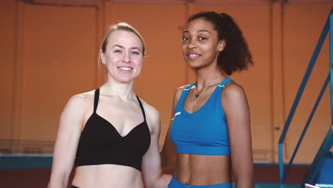 retrato de dos atletas femeninas multiétnicas sonriendo a la cámara mientras están de pie en una instalación deportiva cubierta
