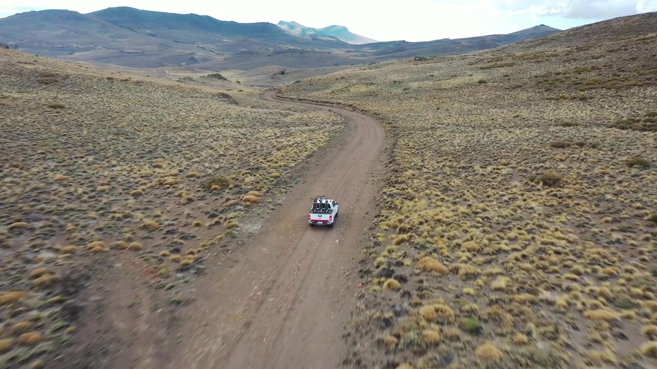 Ripresa con drone dall'alto di un veicolo 4x4 bianco che percorre un sentiero sterrato tortuoso attraverso una vasta valle di montagna a Bariloche, Argentina. Evidenzia viaggi remoti, esplorazione avventurosa e natura selvaggia.