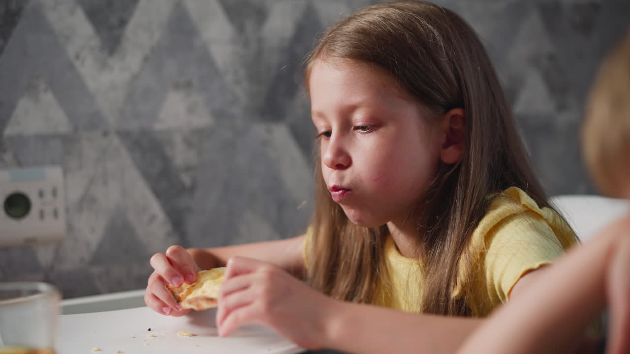 close up of little girl in yellow shirt eating snacks at dining table with transparent glass of juice beside her while person sits next to her slightly out of frame in cozy home environment