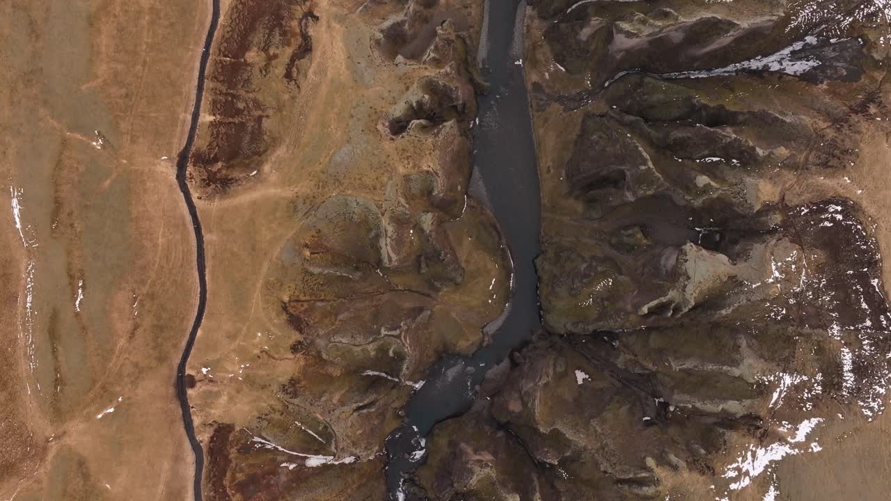 Aerial top-down view of Fjaðrárgljúfur Canyon near Kirkjubæjarklaustur, Iceland. Serpentine river slicing through dramatic ancient rock formations in a barren volcanic landscape.