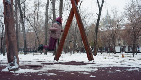 side view of young adult in winter attire swinging on swing while holding chains, wearing maroon trousers and hoodie, with snowy park background and two people running in the distance