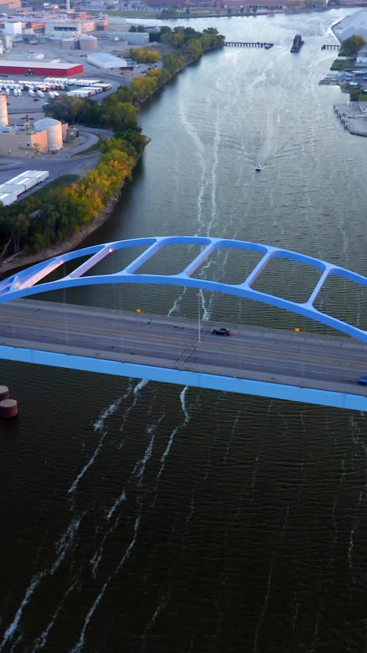 A solitary white car travels across a bright blue arch bridge spanning a calm river beside industrial warehouses and trees, blending motion, geometry, and human engineering in quiet balance
