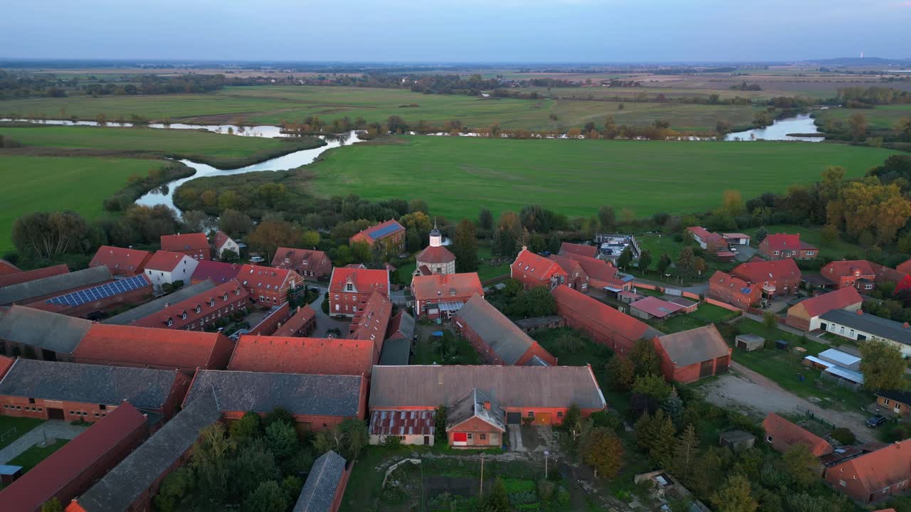 a small village illuminated by golden hour light, casting long shadows over the surrounding fields. Dramatic aerial view flight panorama overview drone