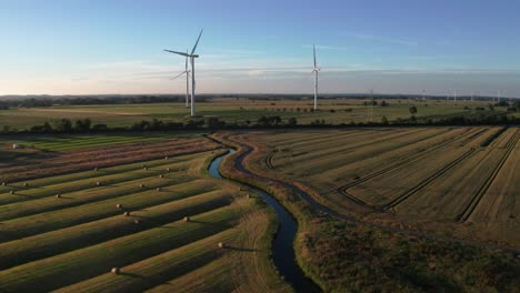 Drone-Disparó-Campo-De-Trigo-Con-Turbinas-Eólicas-A-La-Hora-Dorada-Con-Cielos-Azules-Y-Un-Pequeño-Río