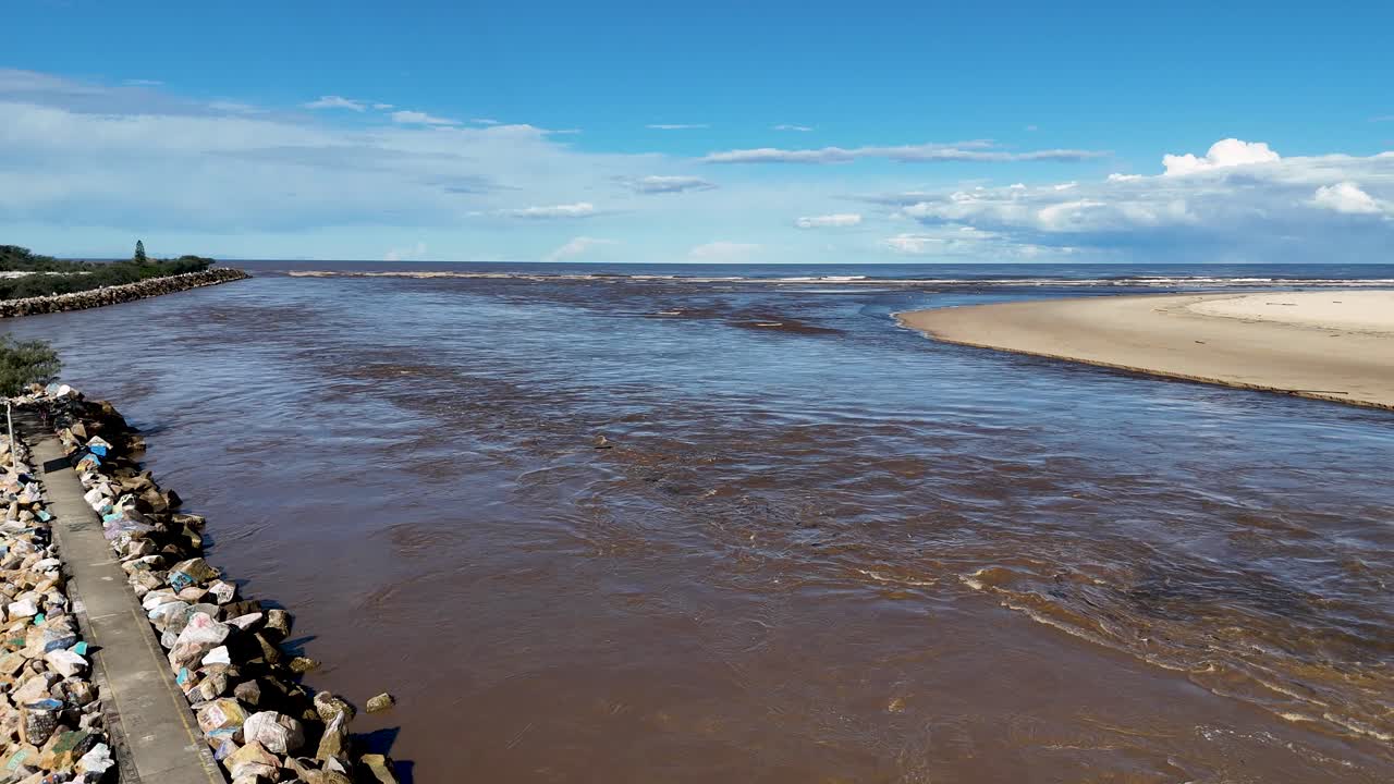 Aerial drone flies over rocky jetty at river mouth, brown water meets sandy beach