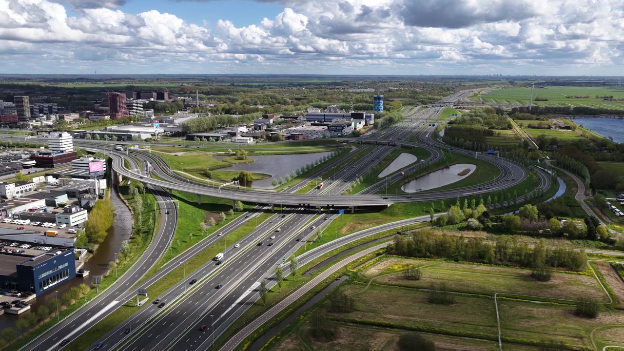 Fugleperspektiv der viser et stort transportknudepunkt i Holland, med køretøjer der bevæger sig langs det komplekse vejnet under en delvist skyet himmel, der forbinder byen med landskabet.