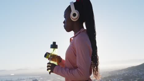 african american woman exercising outdoors wearing wireless headphone drinking water in countryside